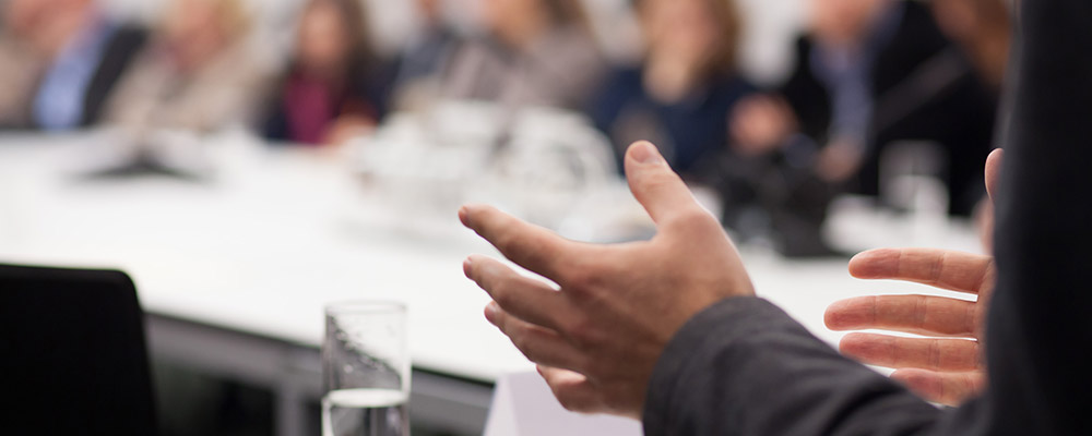 Büroräume, Mitarbeiter laufen durch und sehen beschäftigt aus, eine Konferenz findet statt im Hintergrund.